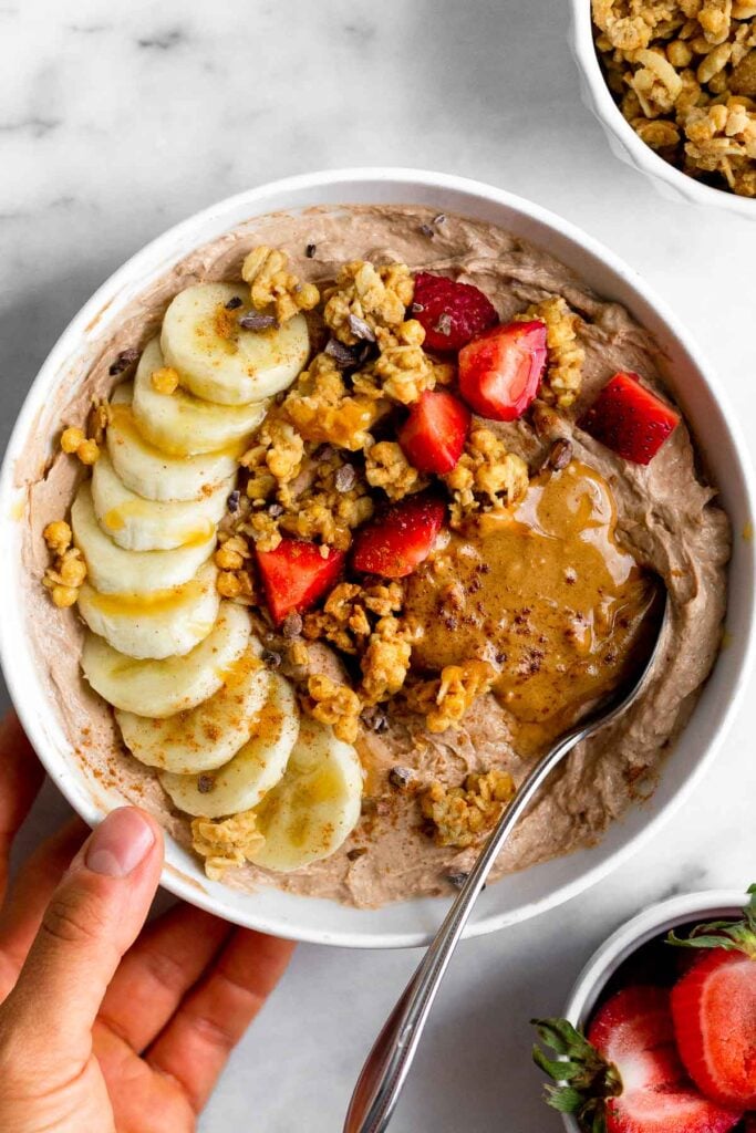 Chocolate protein yogurt in a bowl topped with sliced banana, sliced strawberries, granola, peanut butter, and honey. The bowl is being held up and there is a spoon in it. Underneath it is a bowl of strawberries and another bowl of granola.