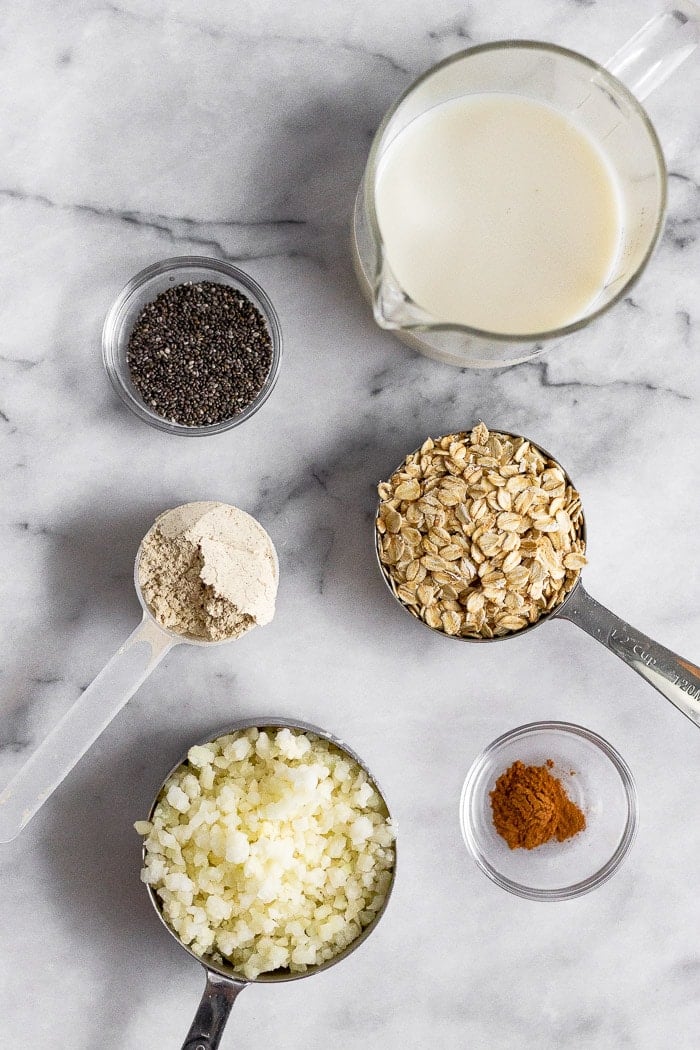 White countertop with a measuring cup of almond milk, a measuring cup of rolled oats, bowl of cinnamon, measuring cup of cauliflower rice, scoop of protein powder, bowl of cinnamon, and bowl of chia seeds.