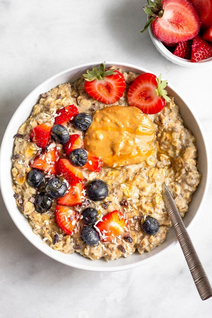 Overhead shot of riced cauliflower oatmeal topped with berries, peanut butter, and honey. A spoon is in the bowl and a small bowl of chopped strawberries is next to the bowl.