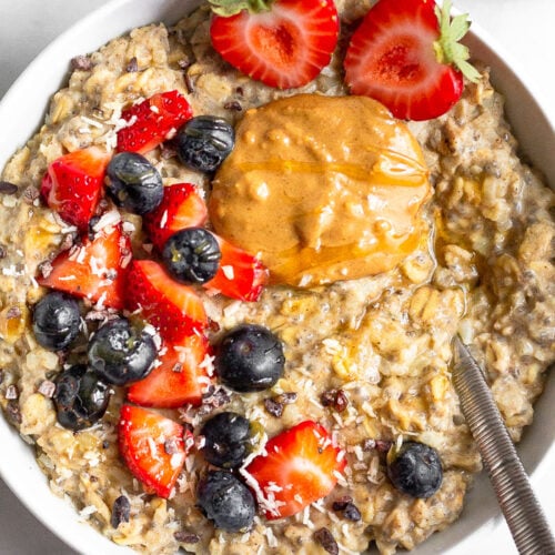 Bowl of cauliflower oatmeal topped with berries and peanut butter with a spoon coming out of it. There is a small bowl of cut up strawberries next to it.