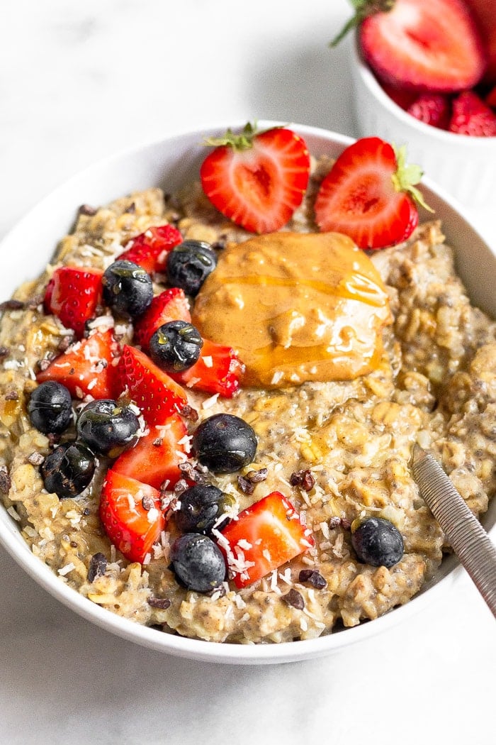 Bowl of cauliflower oats topped with strawberries, blueberries, peanut butter, and honey with a spoon coming out of the bowl. Behind it is a small ramekin of sliced strawberries.