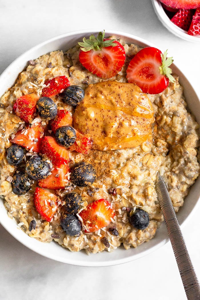 Large bowl of cauliflower protein oatmeal topped with berries, peanut butter, and cinnamon with a spoon in the bowl. Next to it is a small down of diced strawberries.