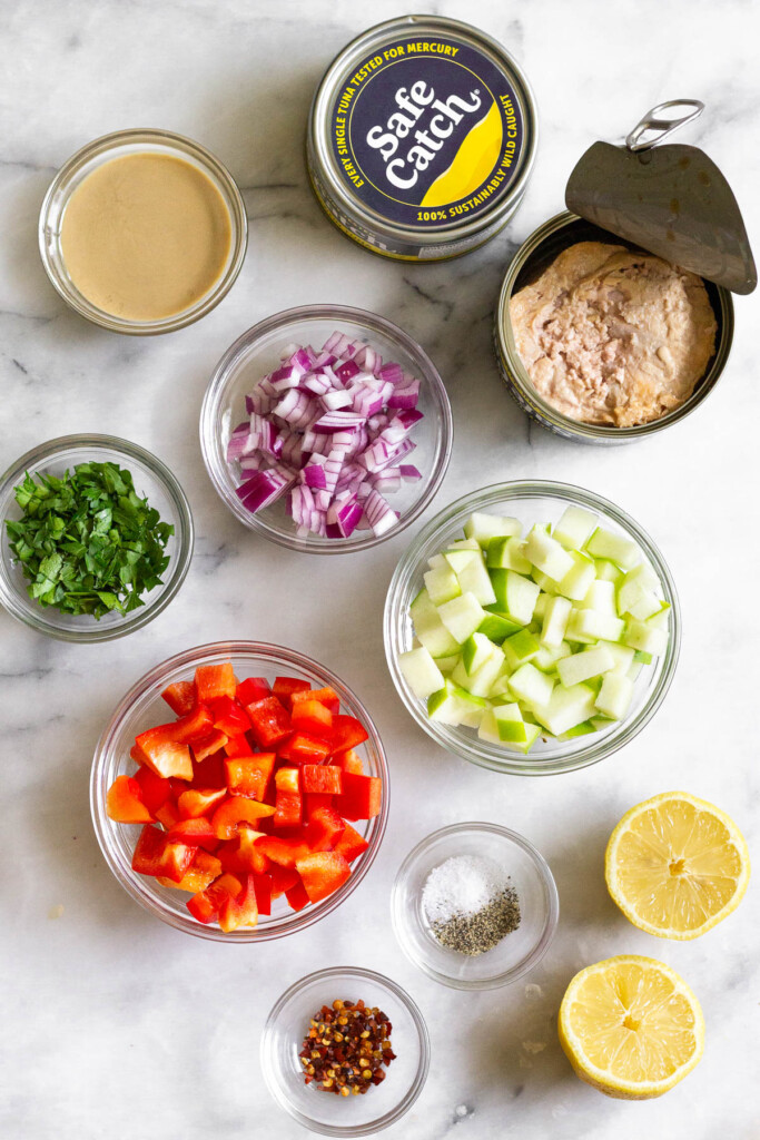 Marble counter with a can of unopened tuna fish, a can of opened tuna fish, a bowl of diced green apple, a lemon that has been cut in half, a small bowl of salt of pepper, a small bowl of red pepper flakes, a bowl of diced red bell pepper, a bowl of diced red onion, a bowl of parsley, and a bowl of tahini on it.