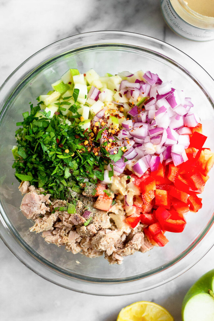 Glass bowl with diced red onion, diced red bell pepper, canned tuna fish, fresh parsley, diced green apple, red pepper flakes, and tahini before it is mixed together. Next to the bowl is a jar of tahini, half a green apple, and half a lemon.