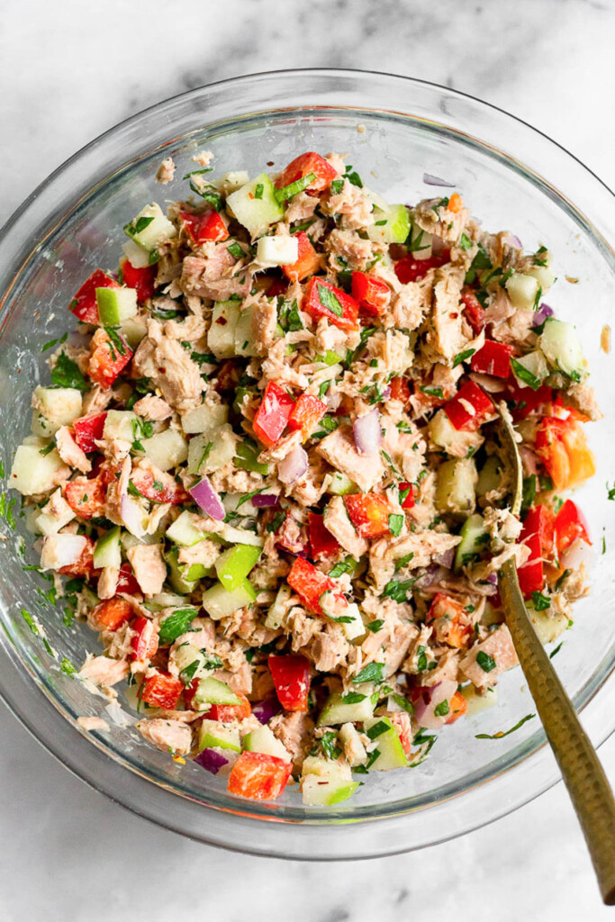 Tuna salad with apples, bell pepper, and herbs tossed in tahini in a glass bowl. There is a spoon in the bowl as well.