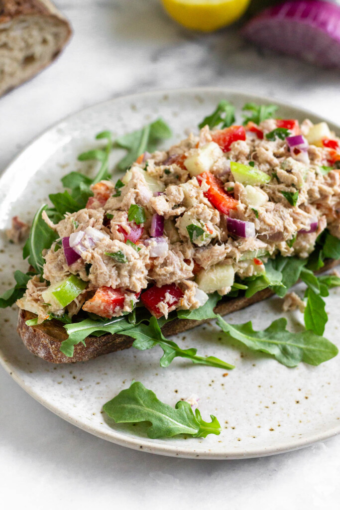 No mayo tuna salad with apples, veggies, and tahini on top of arugula on a piece of toast on a plate. Behind the plate is half a red onion, a lemon, and a loaf of bread that has been cut.