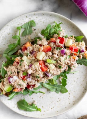 Overhead shot of healthy tuna salad without mayo on top of arugula on a piece of toast on a plate. Next to plate is half a lemon and a red onion.