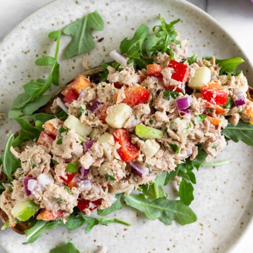 Overhead shot of healthy tuna salad without mayo on top of arugula on a piece of toast on a plate. Next to plate is half a lemon and a red onion.