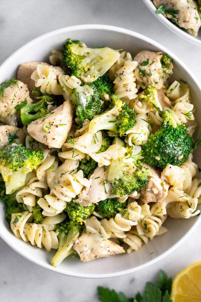 Bowl of chicken and broccoli pasta with ranch dressing. Next to is is another bowl, half a lemon, and some parsley.