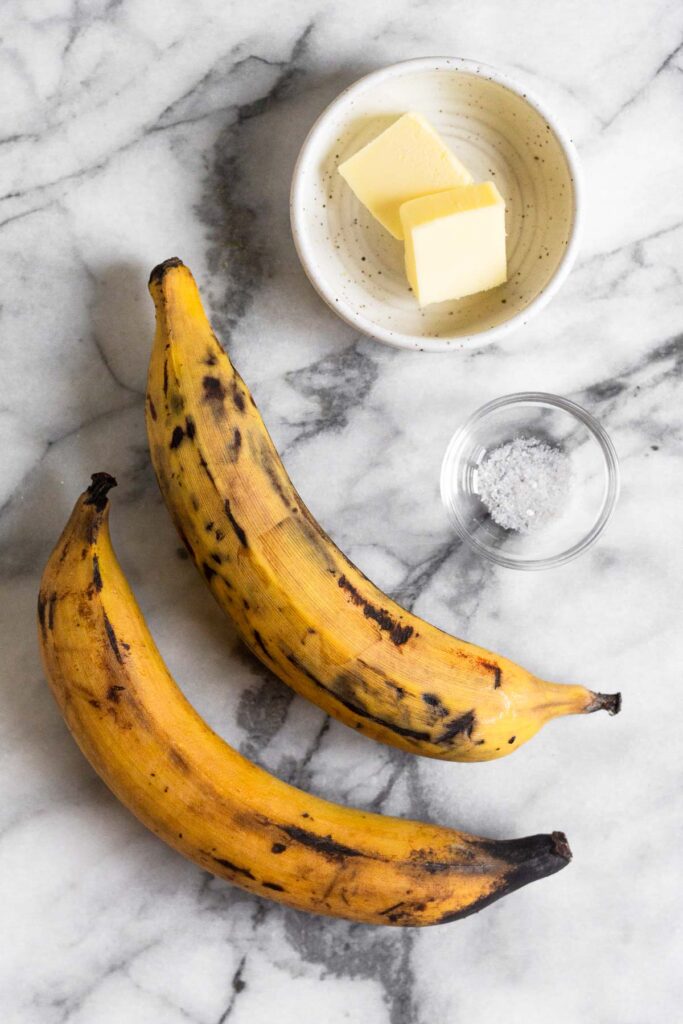 White marble counter with two ripe plantains, a bowl of butter, and a bowl of salt.