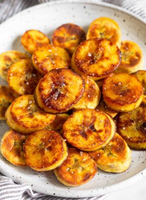A pile of fried sweet plantains on a white plate. They are garnished with flaky salt.