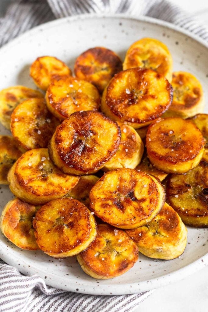 A pile of fried sweet plantains on a white plate. They are garnished with flaky salt.