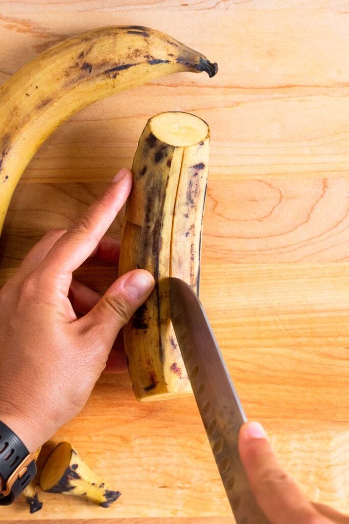 Knife cutting vertically down a ripe plantain on a wooden cutting board. Also on the cutting board is another ripe plantain.