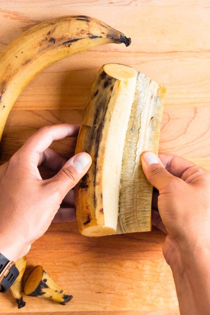 A person peeling a ripe plantain on a wooden cutting board. Another plantain is next to it.