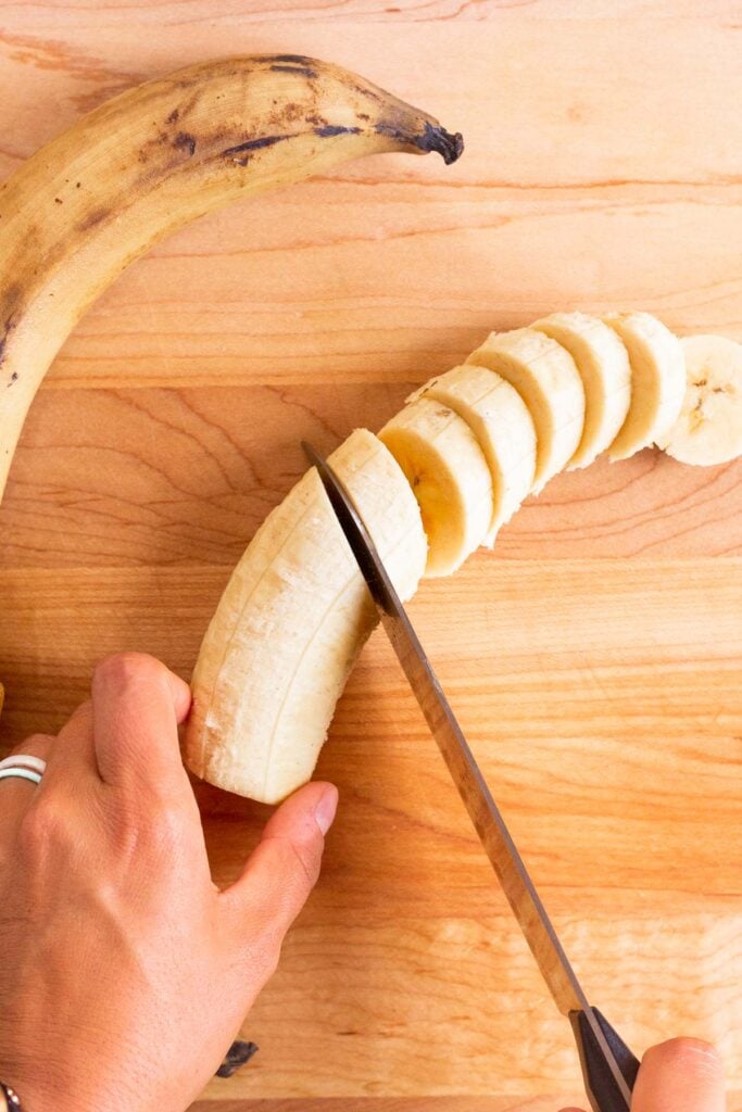 A plantain being sliced into coins on a wooden cutting board. Next to it is whole ripe plantain.