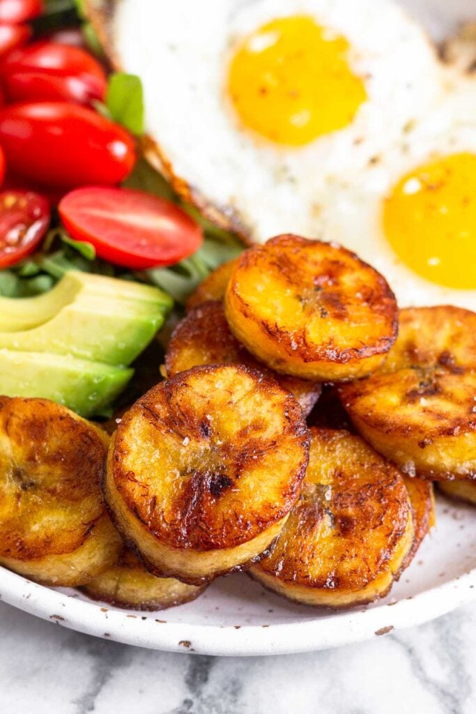 Close up of fried sweet plantains on a plate. Also on the plate is sliced avocado, cherry tomatoes, greens, and fried eggs.
