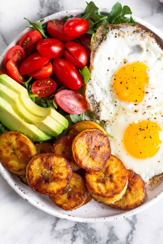 Overhead shot of a plate with two fried eggs, fried plantains, sliced avocado, cherry tomatoes, and greens on it.