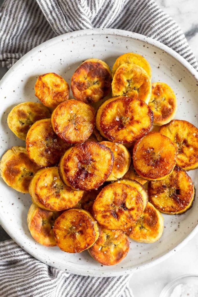 Overhead shot of a pile of platanos maduros garnished with sea salt on a white plate. The plate is sitting on top of a striped towel.