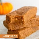 A stack of 3 pumpkin protein bars with a bite taken out of the first one. In the background is a small pumpkin.