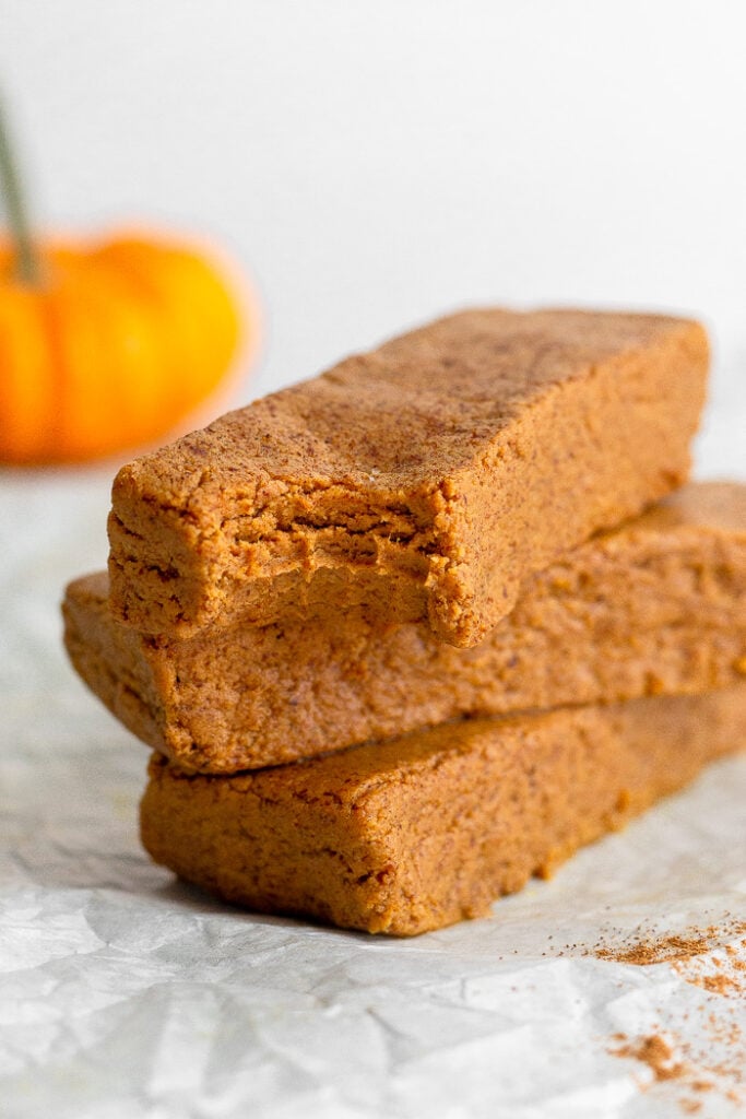 A stack of 3 pumpkin protein bars with a bite taken out of the first one. In the background is a small pumpkin.