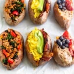 A baking sheet with 6 stuffed breakfast sweet potatoes. Two are stuffed with sausage, peppers, and kale, two are stuffed with bacon, eggs, and avocado, and two are stuffed with fruit and nut butter.