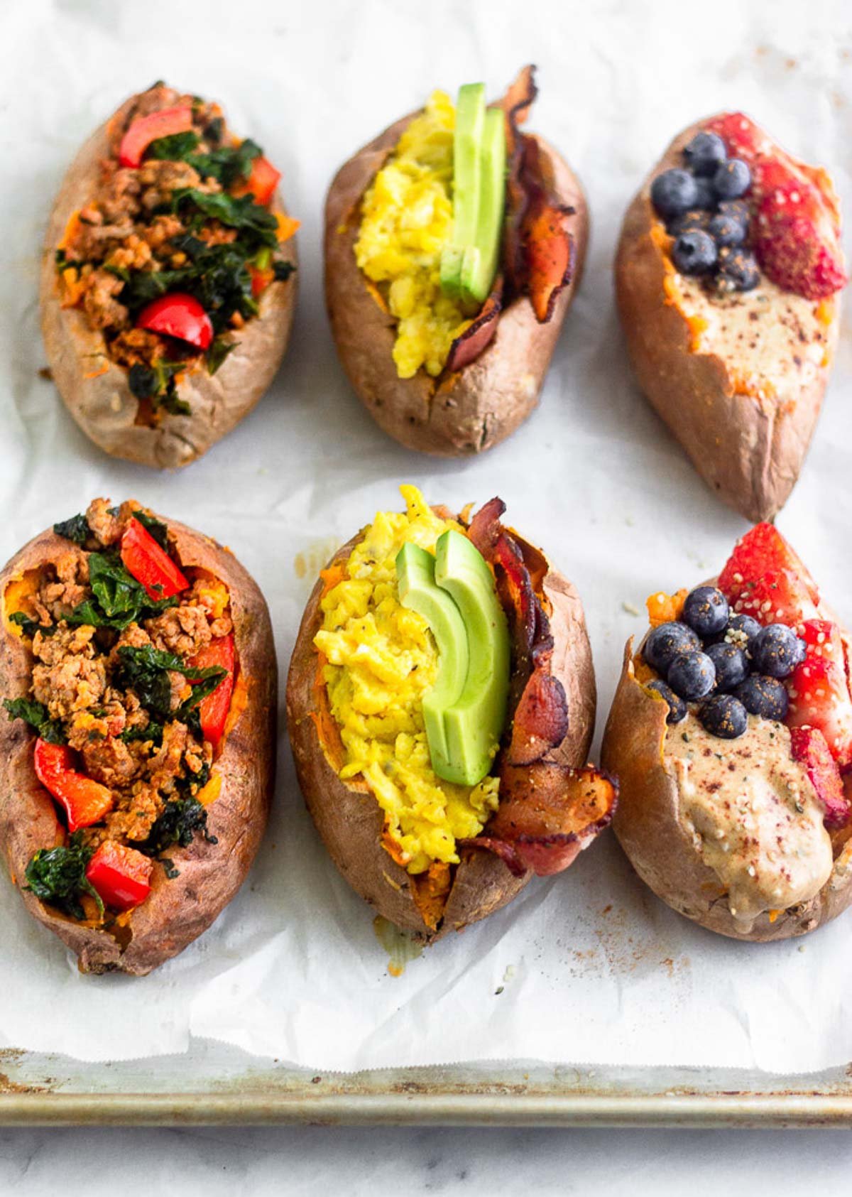 A baking sheet with 6 stuffed breakfast sweet potatoes. Two are stuffed with sausage, peppers, and kale, two are stuffed with bacon, eggs, and avocado, and two are stuffed with fruit and nut butter.