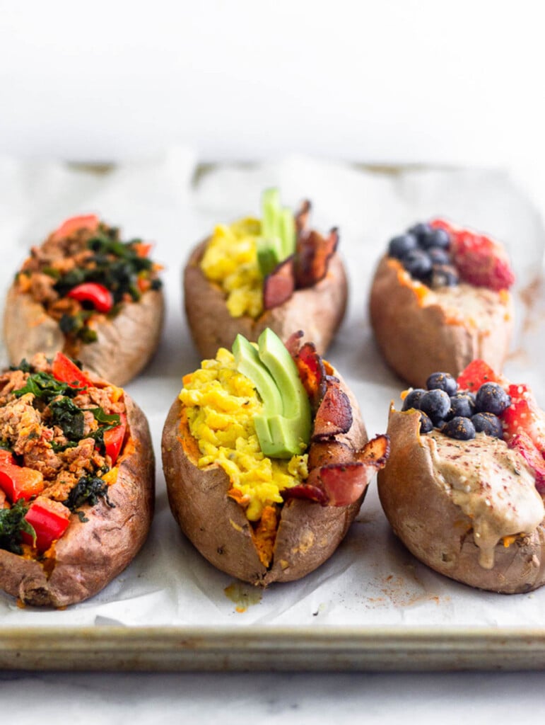 Six breakfast sweet potatoes on a baking sheet. Two are stuffed with sausage, peppers, and kale, two are stuffed with bacon, eggs, and avocado, and two are stuffed with fruit and nut butter.
