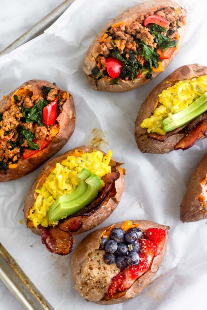 Six different breakfast sweet potatoes on a baking sheet. Two are stuffed fruit and nut butter, two are stuffed with bacon, eggs, and avocado, and two are stuffed with sausage, peppers, and kale.