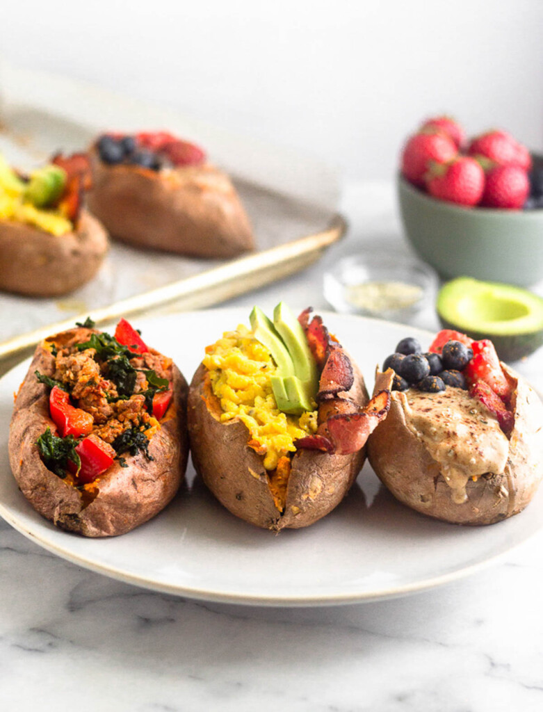 Three different stuffed sweet potato breakfast recipes on a white plate. One sweet potato is stuffed with sausage and veggies, another one is stuffed with scrambled eggs, bacon, and avocado, and the last one is stuffed with fruit and nut butter. Behind them is half an avocado, a bowl of fresh fruit, and a baking tray with more stuffed sweet potatoes.