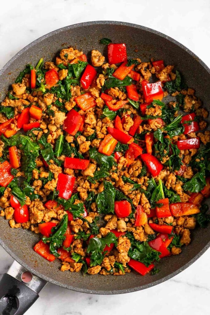 Large skillet filled with a mixture of cooked ground sausage, chopped red bell peppers, and kale.
