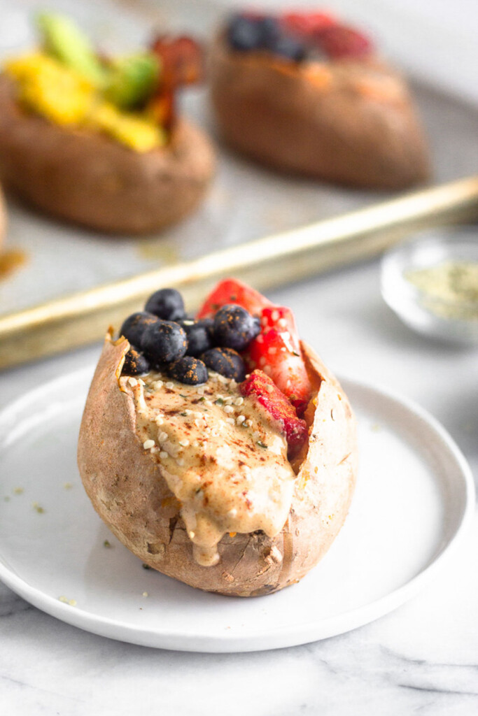 A small plate with a stuffed sweet potato filled with nut butter and berries and topped with cinnamon. Behind it is a baking sheet with different stuffed sweet potatoes.
