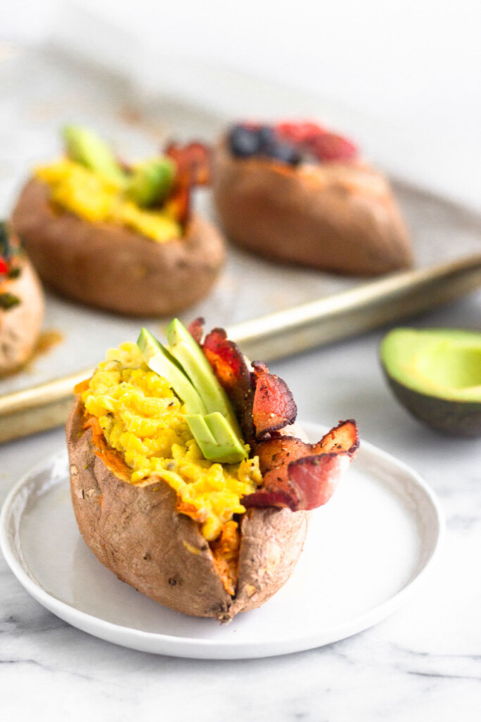 Breakfast sweet potato stuffed with scrambled eggs, bacon, and avocado on a small plate. Behind it is half and avocado and a baking sheet with different stuffed sweet potatoes.