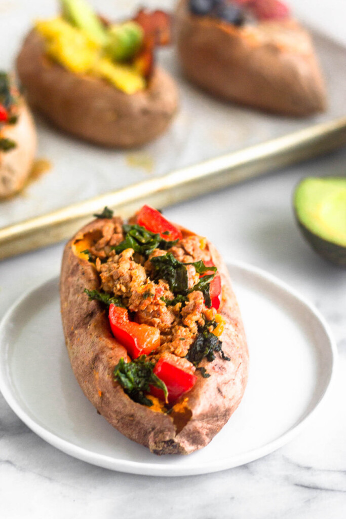 Sweet potato stuffed with a mixture of sausage, red bell peppers, and kale on a small plate. Behind it is half and avocado and a baking sheet with different stuffed sweet potatoes.