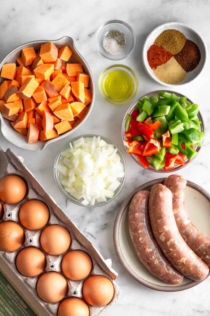 White marble countertop with a small bowl of salt and pepper, a small. bowl of oil, a small bowl of spices, a bowl of diced bell peppers, a plate of 3 sausage links, a carton of eggs, a bowl of diced onions, and a bowl of diced sweet potatoes.