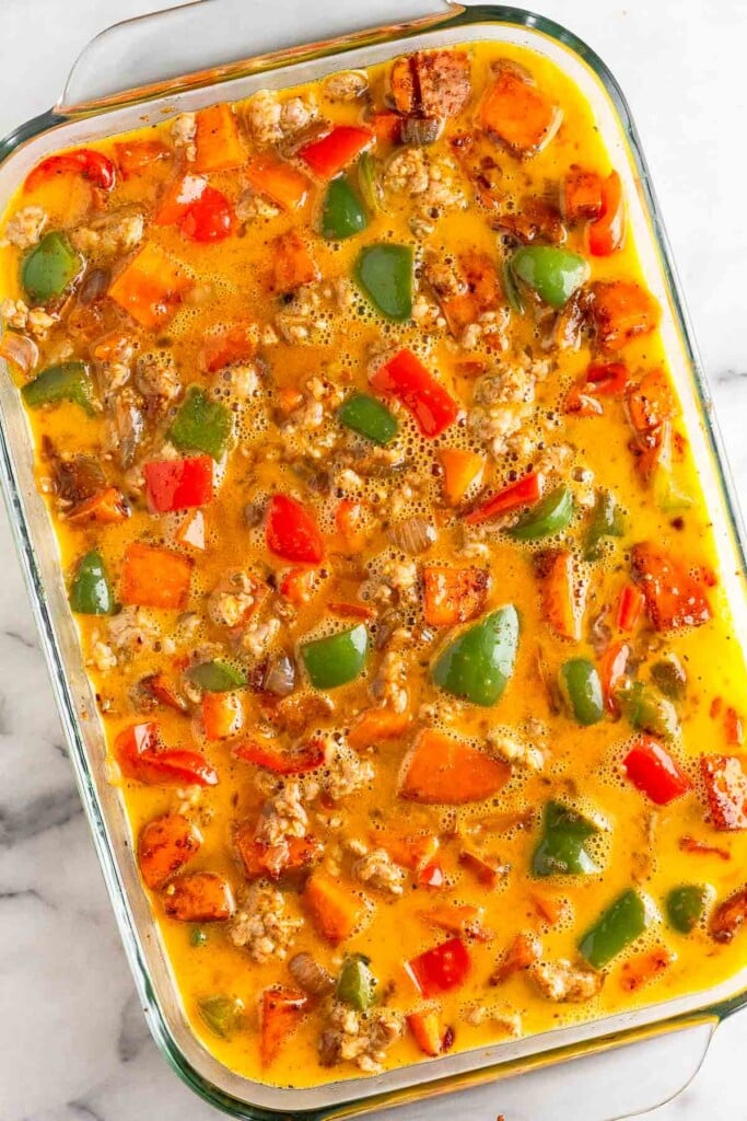 Large glass casserole dish filled with sausage, sweet potatoes, and peppers mixed with whisked eggs.