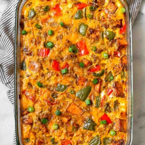 Sweet potato breakfast casserole with sausage and peppers in a large glass casserole dish. The dish is in a striped kitchen towel and next to is is a bowl of chopped green onions and half an avocado.