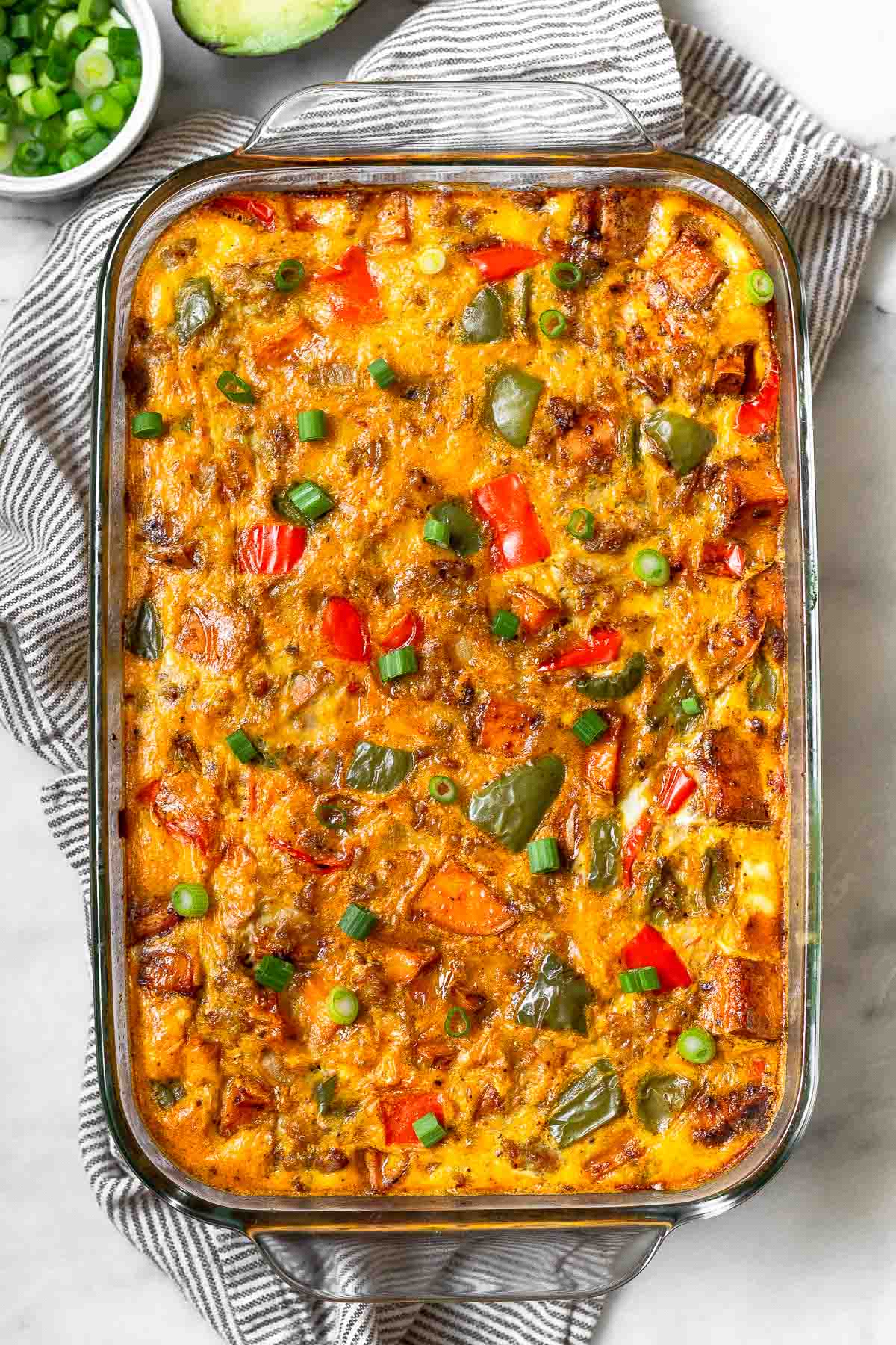 Sweet potato breakfast casserole with sausage and peppers in a large glass casserole dish. The dish is in a striped kitchen towel and next to is is a bowl of chopped green onions and half an avocado.