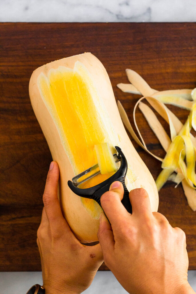 Overhead shot of someone peeling a butternut squash over a wooden cutting board.