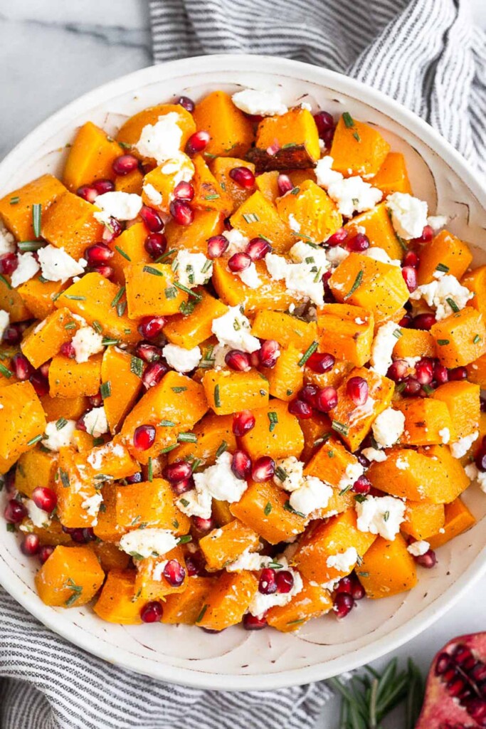 Bowl filled with roasted butternut squash, goat cheese, pomegranates, and rosemary all mixed together. It is sitting on a striped kitchen towel with half a pomegranate and more rosemary next to it.