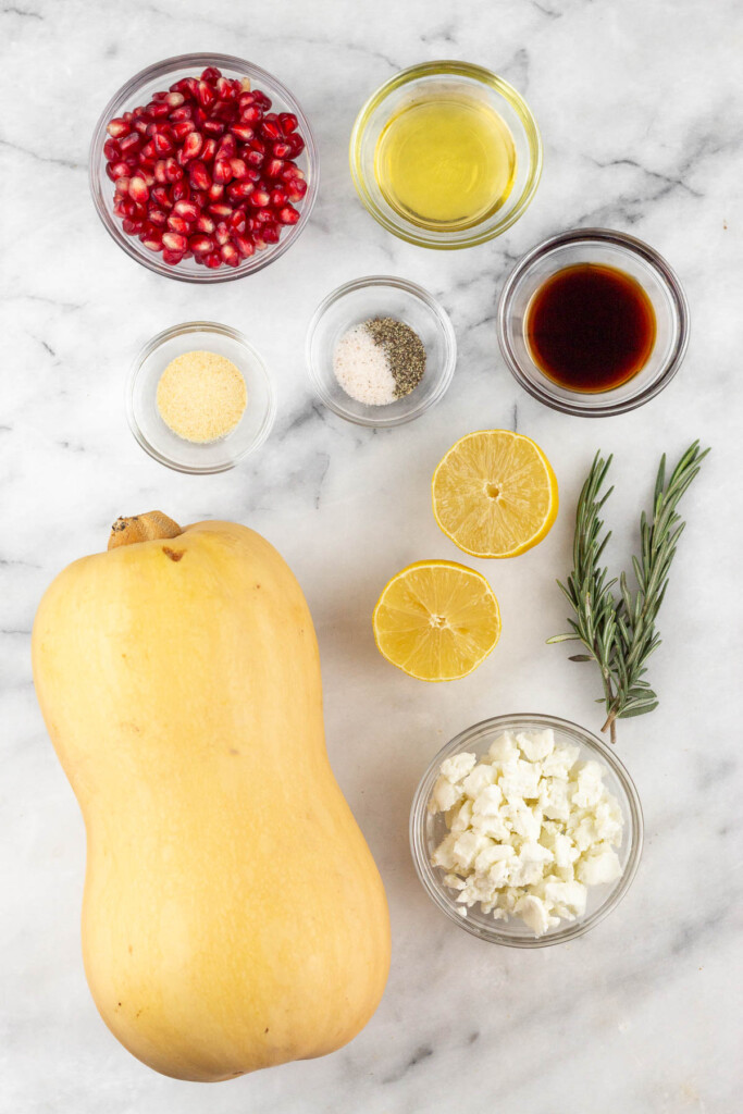 White marble counter with a bowl of olive oil, a bowl of balsamic vinegar, two sprigs of rosemary, a lemon that has been cut in half, a bowl of goat cheese, a whole butternut squash, a bowl of salt and pepper, a bowl of garlic powder, and a bowl of pomegranate seeds.