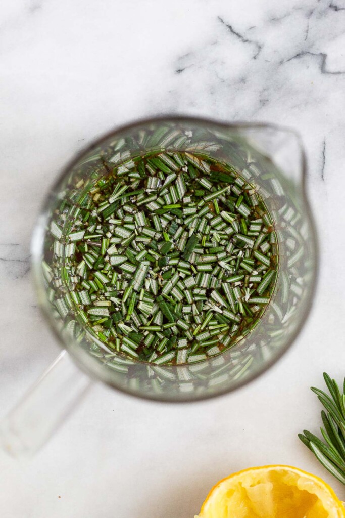 Measuring cup filled with chopped rosemary mixed with lemon juice and balsamic vinegar. Next to it is half a lemon and more rosemary.