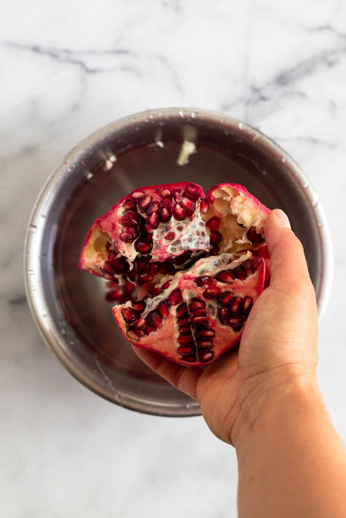 A hand holding half a cut pomegranate over a metal bowl filled with water.