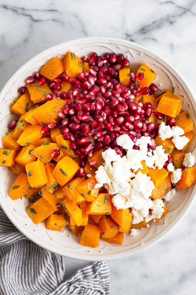 Overhead shot of a large bowl filled with roasted cubed butternut squash, pomegranate arils, and goat cheese before it is mixed together. The butternut squash has some fresh rosemary on it as well.