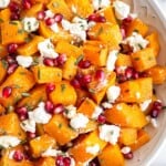 Large white bowl with roasted butternut squash with goat cheese and pomegranates tossed with a rosemary dressing. Next to the bowl is half a pomegranate and some fresh rosemary.