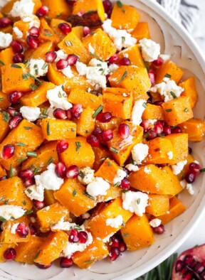 Large white bowl with roasted butternut squash with goat cheese and pomegranates tossed with a rosemary dressing. Next to the bowl is half a pomegranate and some fresh rosemary.