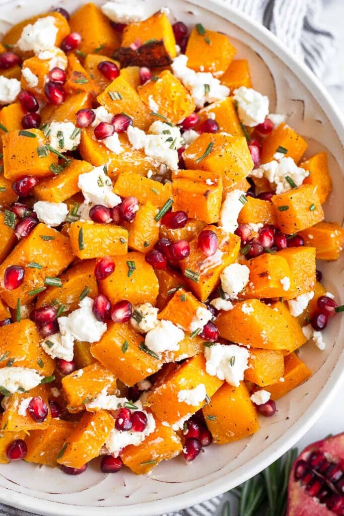 Large white bowl with roasted butternut squash with goat cheese and pomegranates tossed with a rosemary dressing. Next to the bowl is half a pomegranate and some fresh rosemary.