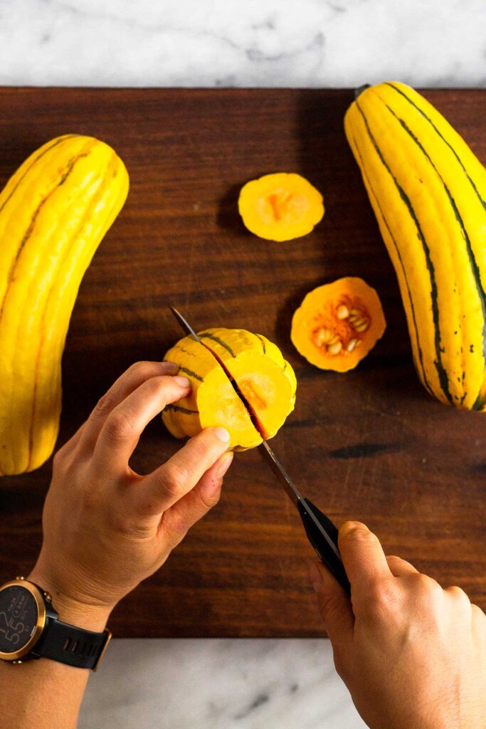 Three delicata squashes on a wooden cutting board with one being cut into lengthwise.