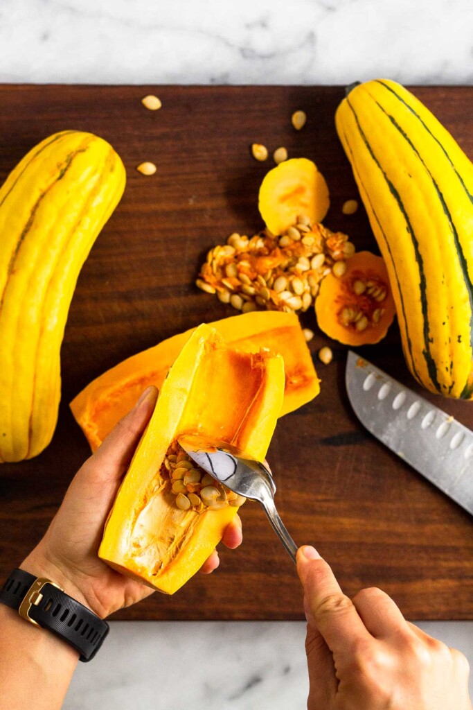 Three delicata squashes on a wooden cutting board. One was halved lengthwise and the seeds are being scooped out.