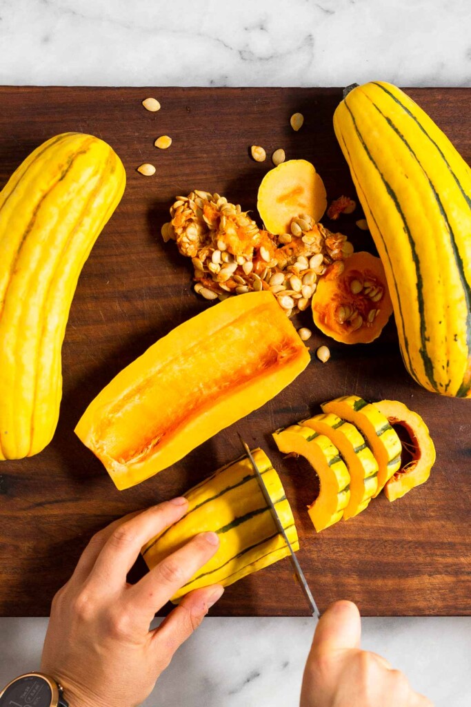 Three delicata squashes on a wooden cutting board. One was halved and one of the halves is being cut into half moon pieces.