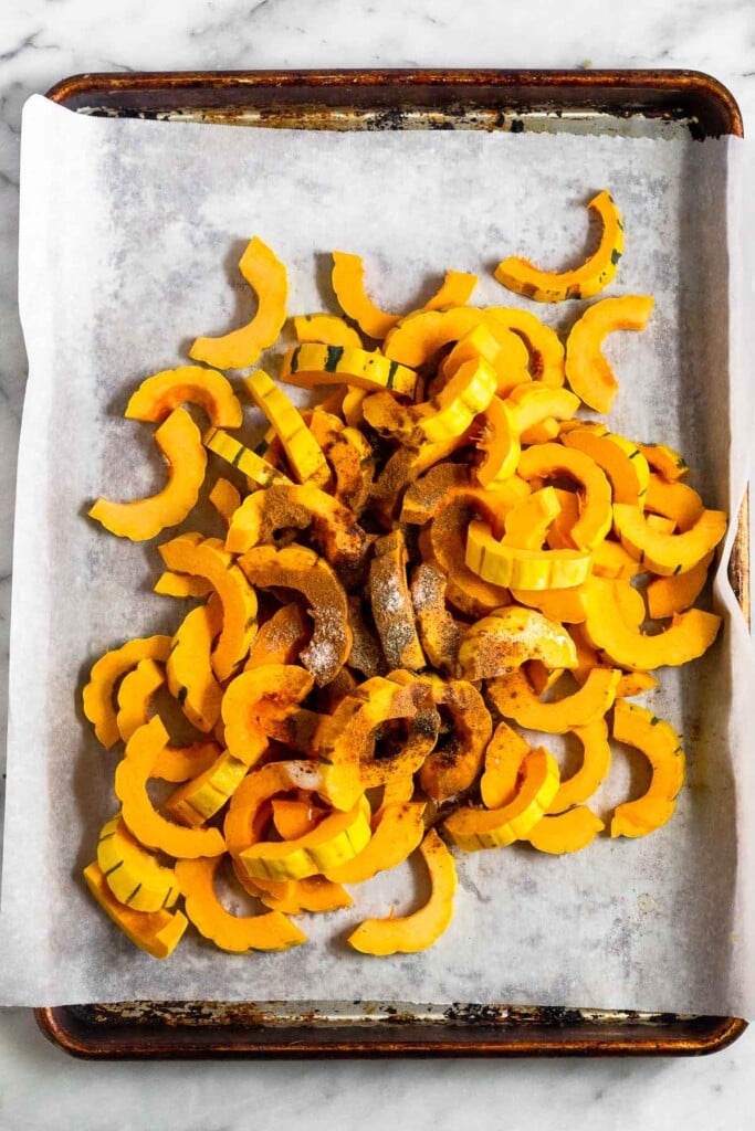 A pile of cut delicata squash a parchment paper lined baking sheet with oil and spices on top of it.